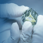 A close-up of gloved hands holding a metallic medical device, likely a stent or heart valve, against a blurred background.